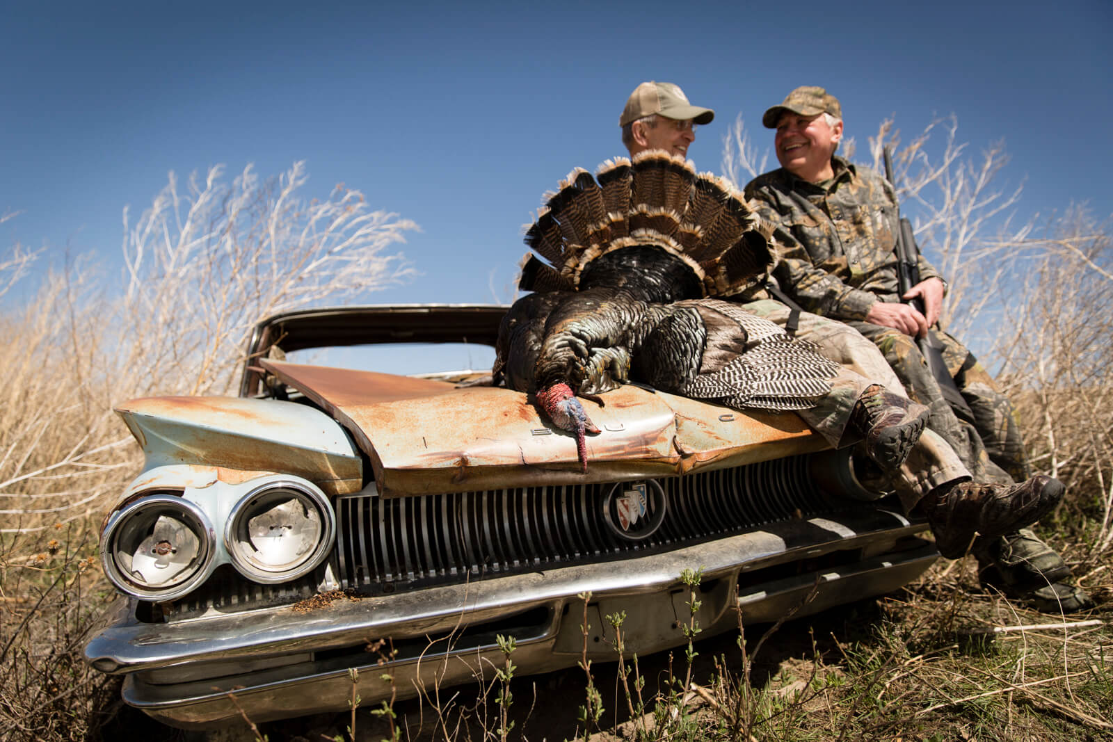 Two hunters and a successfully hunted turkey pose on a rusted car in a field