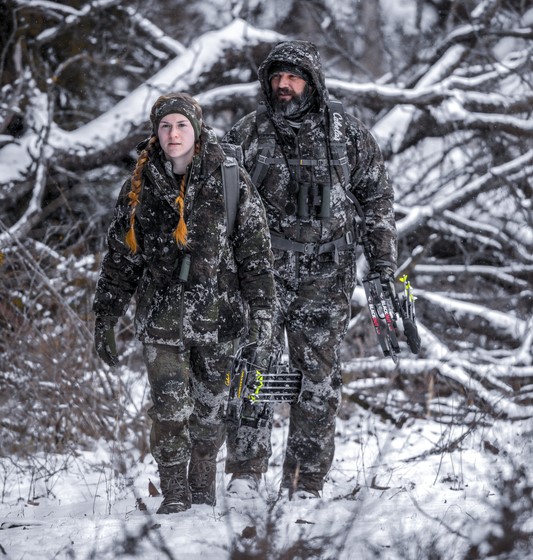 two hunters with compound bows trekking through snow-covered woods