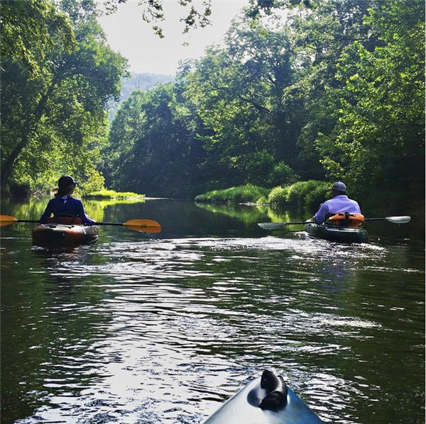 Two kayakers floating the current river through a green canopy of trees on calm water