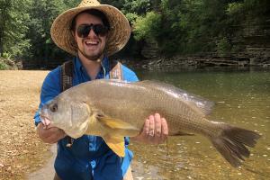Large drum fish held by angler wearing a straw sun hat and blue shirt at waters edge.