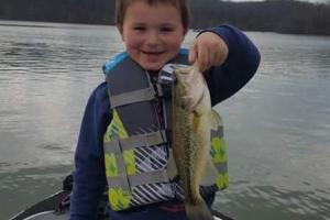 Boy standing in the front of a bass boat on an overcast day hold up a bass