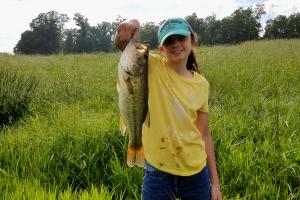 Girl angler standing in a field holding up a large bass