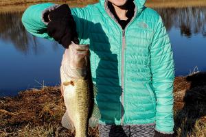 Girl angler standing next to a pond holdingup a largemouth bass