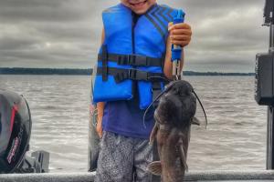 Young boy standing an a boat seat holding a catfish